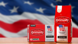 Packages of Community Coffee American Classic medium roast sit on a wooden counter, with a waving American flag filling the background.