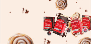 Packages of Community Coffee Chocolate Lava Cake and Cinnamon Roll flavored coffees float among cinnamon rolls and bits of chocolate cake on a cream-colored background.