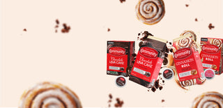 Packages of Community Coffee Chocolate Lava Cake and Cinnamon Roll flavored coffees float among cinnamon rolls and bits of chocolate cake on a cream-colored background.
