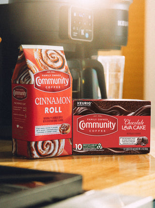Packages of Community Coffee Cinnamon Roll and Chocolate Lava Cake flavored coffees sit on a kitchen counter, in front of a coffee maker, in soft morning light.
