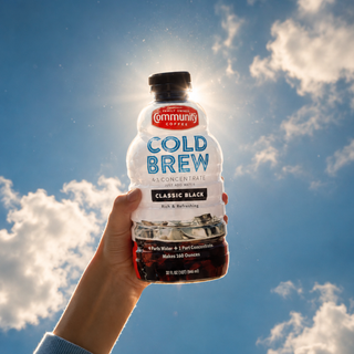 A bottle of Community Coffee Classic Black Cold Brew Concentrate, being held up against a blue sky with clouds and rays of sunshine.
