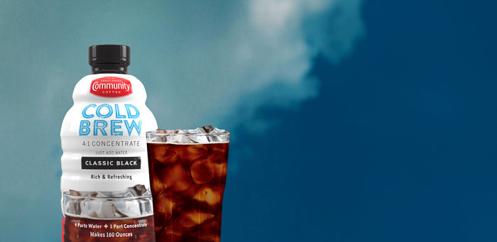 A bottle of Community Coffee Classic Black Cold Brew Concentrate next to a glass of iced coffee, standing against a blue sky with clouds.