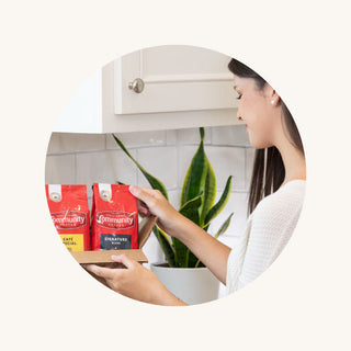 A woman pulls a bag of ground Community Coffee Signature Blend dark roast out of a box.