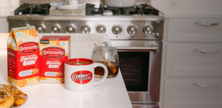 Packages of Community Coffee Glazed Honey Bun flavored coffee sit on a white kitchen counter, next to a mug of coffee and jar of honey.