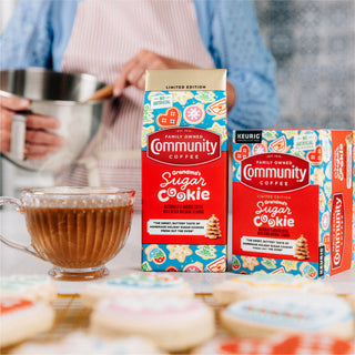 Packages of Community Coffee Grandma's Sugar Cookie flavored coffees sit side-by-side on a kitchen counter, next to a coffee mug, with a person holding a mixing bowl in the background, and cookies in the foreground.