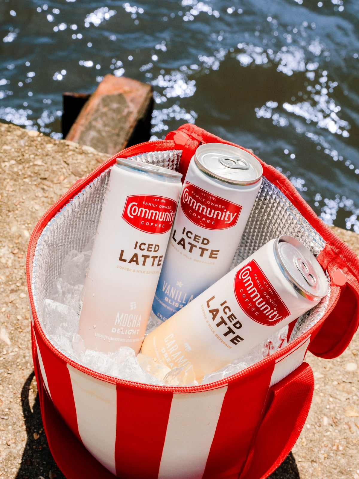 Three Community Coffee Iced Lattes rest in ice in a cooler by water.