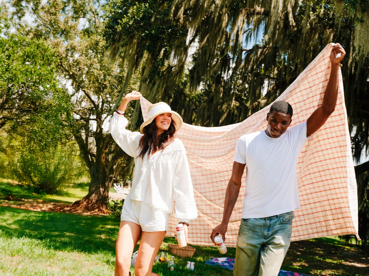 Two young people walk in a wooded area and hold up a picnic blanket while holding Community Coffee Iced Latte cans.
