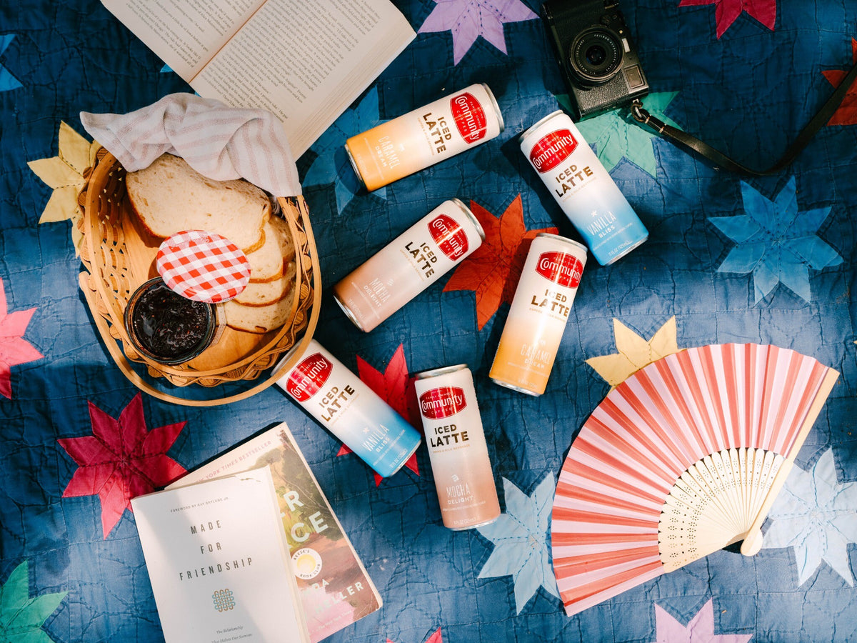 Six Community Coffee Iced Latte cans lie flat on a picnic blanket, next to books and a picnic basket.