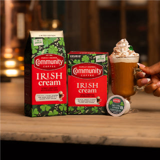 Packages of Community Coffee Irish Cream flavored coffee sit on a wood countertop, next to a glass of rich brown coffee topped with whipped cream.
