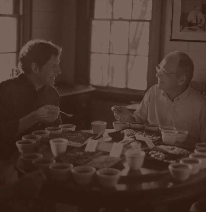 Fourth-generation Community Coffee owner Matt Saurage and third-generation owner Norman Saurage the Third share a smile while testing coffees around a table.
