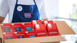 A person wearing a blue USO apron carries a box full of Community Coffee packages.