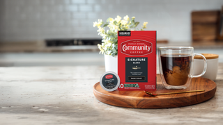 A ten-count box of Community Coffee Signature Blend dark roast K-Cups, sitting next to a creamed cup of coffee and stirrer spoon on a countertop.