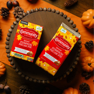 Packages of Community Coffee Spiced Pumpkin Pecan Pie flavored coffee lie on a fall-themed table, featuring plastic pinecones and pumpkins.