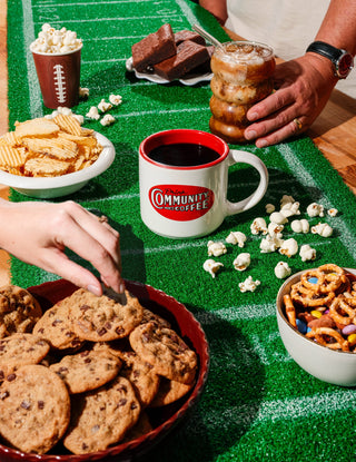 A white coffee mug with the vintage Community Coffee logo sits on a turf table runner, surrounded by game-day snacks.